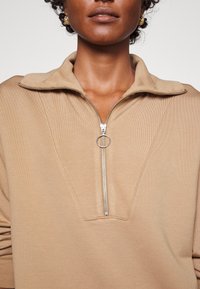 Beige Zip-up-Fleece-Sweatshirt mit Stehkragen, ausgestattet mit einem silbernen Reißverschluss und einem Ringzug, strukturiertem Stoff und schrägen Nahtdetails.