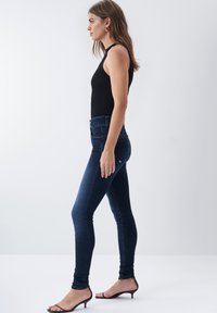Jeans skinny azul oscuro de alta cintura, con una textura de denim suave. Llevados con una blusa sin mangas negra y sandalias de tacón negras.