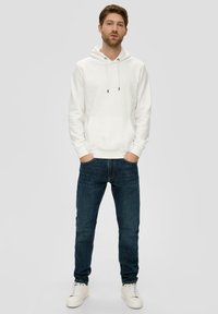 Haut à capuche en coton blanc avec une poche avant et un cordon de serrage, associé à un jean bleu foncé et des baskets blanches. Le modèle se tient droit.