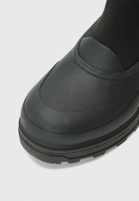 Chaussure noire imperméable avec une finition lisse et mate, dotée d'un bout arrondi et d'une semelle en caoutchouc texturé, garantissant adhérence et stabilité.