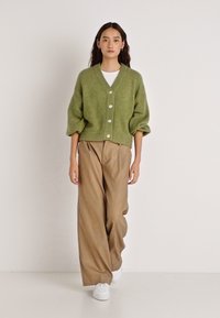 Πράσινη μάλλινη ζακέτα με λαιμόκοψη σε σχήμα V, τρία μεγάλα κουμπιά και φουσκωτά μανίκια, σε συνδυασμό με χαλαρά καφέ παντελόνια και λευκά αθλητικά παπούτσια.