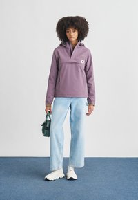 Ljuslila pullover-jacka med framficka, huva och elastiska muddar, kombinerad med ljusblå jeans med vida ben och vita sneakers.