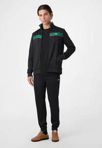 Zwarte jas met rits en groene en rode strepen, gecombineerd met zwarte broek en bruine sneakers. Voorzien van een logo op de borst en de broek.