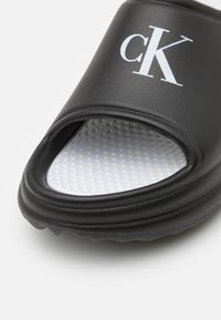 Sandale noire à semelle plate avec une tige texturée, logo blanc "cK", design ouvert à l'avant, et une semelle intérieure blanche, rembourrée et à pois.
