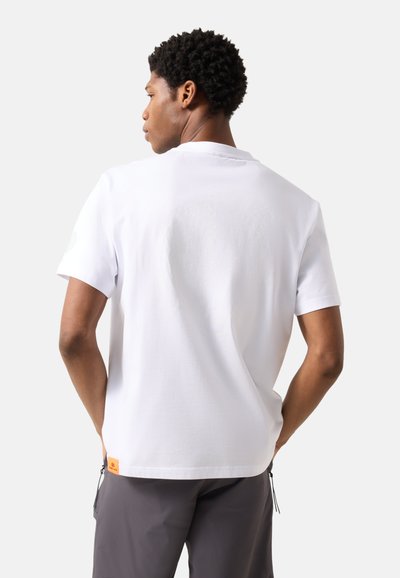 Homme de dos portant un t-shirt blanc uni à manches courtes et un pantalon gris foncé, sur un fond gris clair.