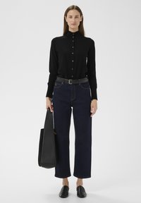 Fekete gombos ing magas gallérral, sötétkék bő szárú farmerrel párosítva. Fekete bőrtáskával és fekete belebújós cipővel viseli.