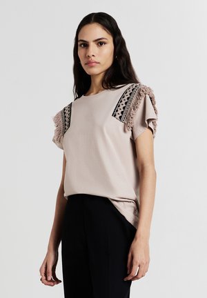 T-shirt rosa chiaro con maniche corte e frange, dettagli in pizzo nero sulle spalle, abbinato a pantaloni scuri, presentato su uno sfondo neutro.