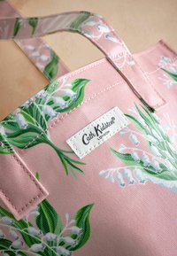 Rozā auduma iepirkumu soma ar ziedu rakstu, kurā ir zaļas lapas un baltas puķes, ar baltu etiķeti, kurā ir uzraksts "Cath Kidston London."