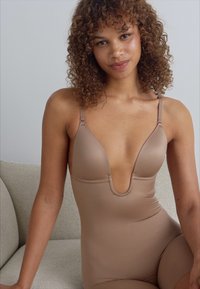 Huiskleurige bodysuit, lage plunge-halslijn, dunne verstelbare bandjes, zachte stof, getailleerde silhouet, gladde textuur. Model zittend op een bank.