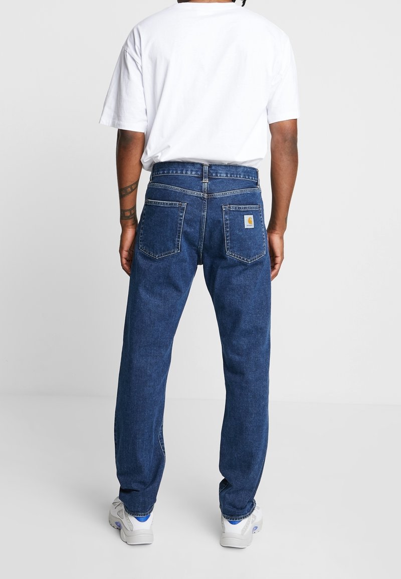 Jeans de mezclilla azul oscuro con un corte relajado, con bolsillos traseros y un pequeño parche de logo. Combinados con una camiseta blanca lisa y zapatillas.