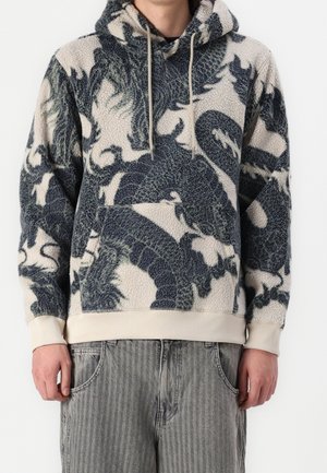 Personne portant un sweat à capuche en polaire avec des motifs de dragon sombres et une bordure beige, associé à un jean rayé gris.