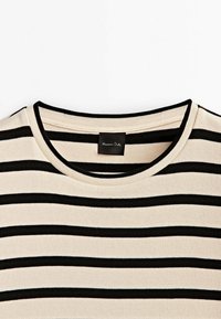 T-shirt en maille beige et noire à rayures horizontales avec col rond, montrant la partie supérieure incluant l’étiquette de la marque à l’intérieur du col.
