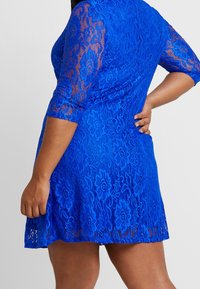 Krótka niebieska koronkowa sukienka z rękawami o długości trzech czwarte, w kwiatowy wzór i rozkloszowanym kroju. Posiada dopasowany gorset i lekko przezroczystą teksturę.