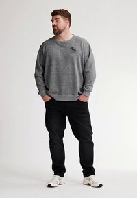 Grå fleece-tröja med rund halsringning och broderad logotyp, kombinerad med svarta jeans och vita sneakers. Modellen står avslappnat med händerna i fickorna.