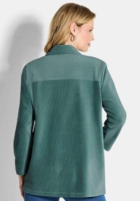 Türkise Cordjacke mit Kragen, langen Ärmeln und lockerer Passform. Die Textur hat vertikale Rippungen und ein glattes Rückenteil.