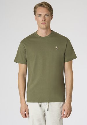 Giovane uomo con capelli corti castano chiaro che indossa una maglietta verde oliva a maniche corte e pantaloni di colore chiaro, in piedi davanti a uno sfondo semplice.