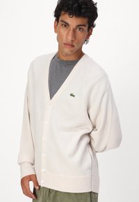 Hvit cardigan laget av et mykt, teksturert stoff med dyp V-hals, frontknapper og en liten grønn logoaksel på brystet.
