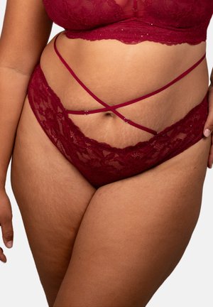 Curvy woman modeling red lace lingerie set with crisscross straps on hips, showing midsection and upper thighs against plain background.