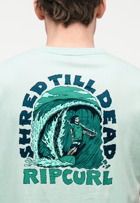 Πίσω μέρος ενός γαλάζιου πουκάμισου με γραφικό σχεδιασμό ενός σκελετού που κάνει σέρφινγκ σε μεγάλο κύμα, περιβαλλόμενο από τις λέξεις «Shred Till Dead Ripcurl.»