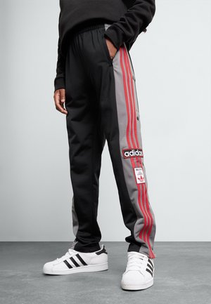 Person wearing black and gray Adidas track pants with red stripes and white Adidas sneakers standing on gray floor.