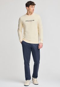 Sudadera de color crema claro con "POLO CLUB ESTABLECIDO 2013" impreso en negro, combinada con pantalones navy y zapatillas beige.