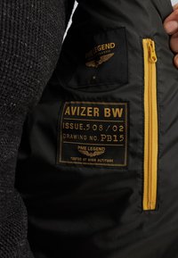 Čierna bunda s interiérom, ktorý obsahuje žltý zips a látkovú nálepku s embosovaným textom. Významné detaily zahŕňajú logo PME Legend a veľkosť "M."