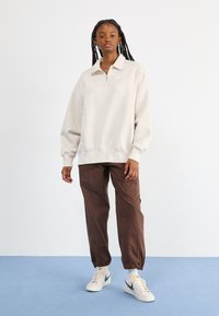 Ανοιχτό καφέ ζακέτα με μισό φερμουάρ, με ραμμένα μανίκια και τελείωμα. Συνδυασμένη με καφέ tapered παντελόνι και λευκά αθλητικά παπούτσια με τόνους του τιρκουάζ.