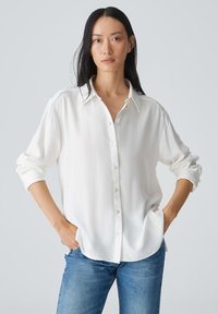 Chemise blanche à manches longues en tissu lisse, avec un col et des manches retroussées, associée à un jean en denim bleu.