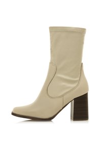 Botín beige hasta el tobillo con un acabado suave, punta afilada y un tacón cuadrado alto y estructurado. Presenta un panel lateral para facilitar el uso.