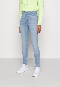 Jeans ajustados de denim en azul claro, con una cintura alta, un sutil desvanecimiento y cinco bolsillos, combinados con una sudadera verde brillante y zapatillas blancas.