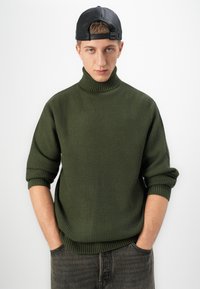 Junger Mann mit den Händen in den Taschen, der einen dunkelgrünen gestrickten Rollkragenpullover und eine rückwärts tragende schwarze Baseballmütze vor einem weißen Hintergrund trägt.