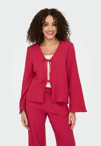 Giacca leggera color cremisi con scollatura a V e chiusure con cordino, abbinata a pantaloni larghi coordinati in un tessuto testurizzato.