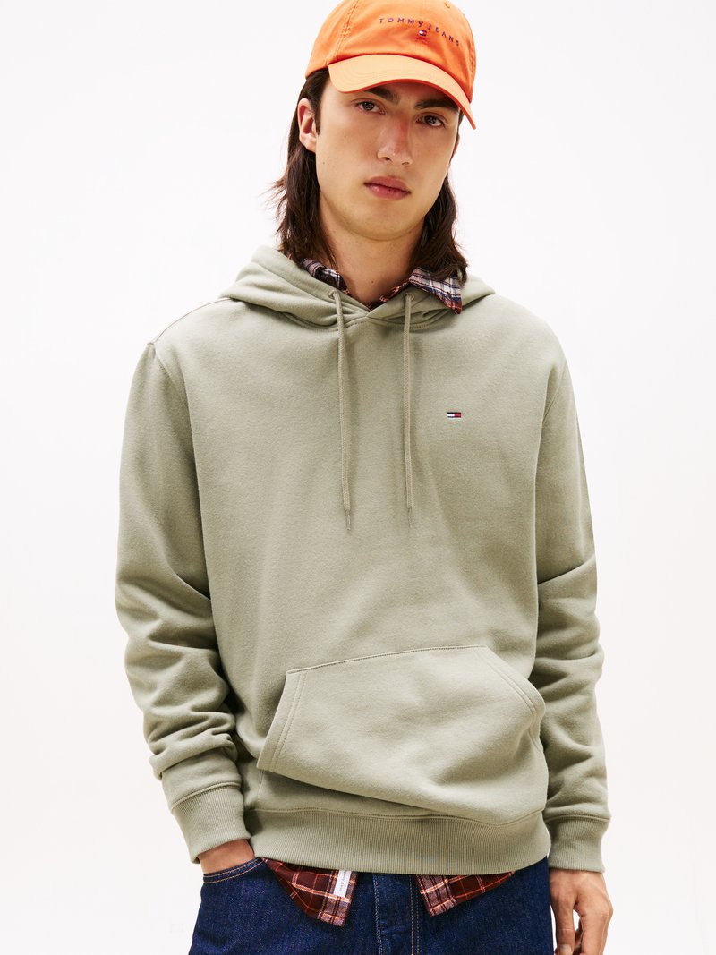 Ljus olivgrön hoodie med en framficka och snoddhuva, bärs över en rutig skjorta, kombinerad med en lysande orange keps.