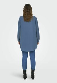Camisa azul de manga larga con dobladillo redondeado, combinada con unos vaqueros ajustados azules y botas negras. Tejido suave y diseño casual.