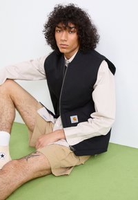 Schwarze Nylonweste mit Reißverschluss vorne, Seitentaschen und gesticktem Logo, über einem hellbeigen Hemd und khakifarbenen Shorts getragen. Grüne Fläche darunter.