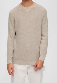 Maglione beige lavorato a maglia con un motivo strutturato, collo rotondo, maniche lunghe e polsini e orlo a costine. Abbinato a pantaloni chiari.