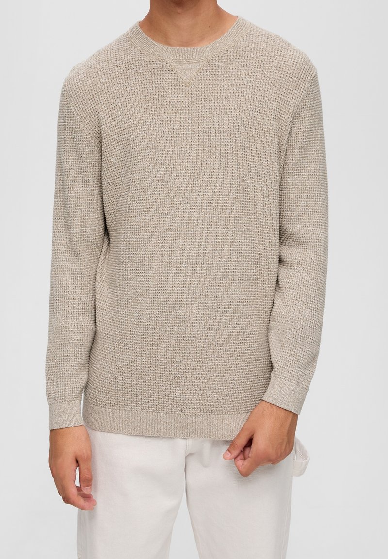 Maglione beige lavorato a maglia con un motivo strutturato, collo rotondo, maniche lunghe e polsini e orlo a costine. Abbinato a pantaloni chiari.