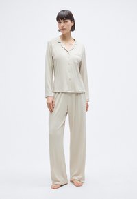 Σετ μπεζ πυτζάμες με πουκάμισο μακρυμάνικο με κουμπιά και τσέπη, καθώς και φαρδιά παντελόνια. Κατασκευασμένο από λεία, ελαφριά ύφασμα.