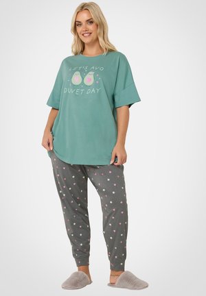 Kvinde smilende, iført en teal oversized T-shirt med teksten "Let's Avo Duvet Day", grå pyjamasbukser med stjerner og hjerter og grå bløde tøfler.