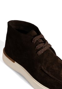 Donkerbruin suède enkelboots met gestructureerde brown veters en een dikke witte zool, weergegeven in een close-up schuin zicht.