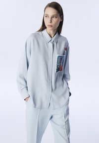 Camicia azzurro chiaro con bottoni, dotata di tasca sul petto con una stampa floreale colorata. Progettata con una vestibilità comoda e maniche lunghe.