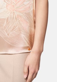 Hellrosa Bluse mit floralem Muster, durchscheinendem Satin-Stoff und einem kleinen orangen Knopfdetail am Saum, kombiniert mit einem beigen, strukturierten Unterteil.