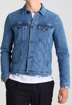 Man wearing a blue denim jacket with buttoned chest pockets and frayed hem over a white shirt and black pants.