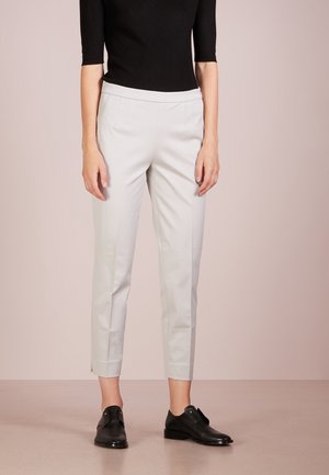 Vrouw draagt lichtgrijze, op maat gemaakte enkelhoge broek, zwart top met halflange mouwen en zwarte leren instapschoenen tegen een effen achtergrond.