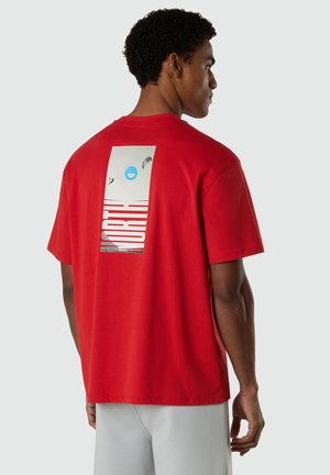 Mann trägt ein rotes T-Shirt mit einem rechteckigen Motiv und dem Wort "NORTH", das vertikal auf dem Rücken gedruckt ist, kombiniert mit hellgrauen Hosen.