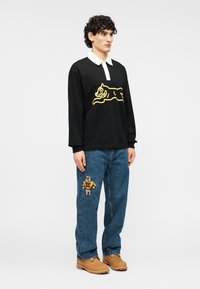 Μαύρο πουκάμισο με μακριά μανίκια και κίτρινο γραφικό γάτας, λευκό γιακά; μπλε τζιν με κεντημένο πατς αρκούδας; καφέ μποτάκια. Λείο ύφασμα και άνετη γραμμή.