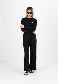 Black long-sleeve top and wide-leg pants, ribbed texture, small logo on the chest, paired with black flip-flops and sunglasses.