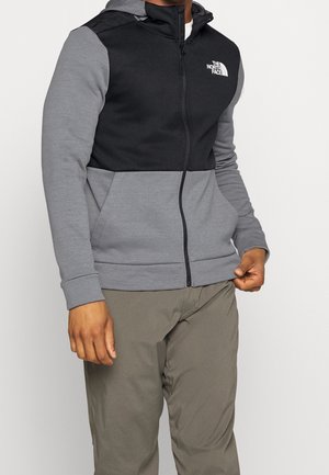 Mann trägt einen schwarz-grauen Kapuzenpullover mit Reißverschluss und dem The North Face-Logo sowie olivgrüne Hose, steht vor einem schlichten hellen Hintergrund.