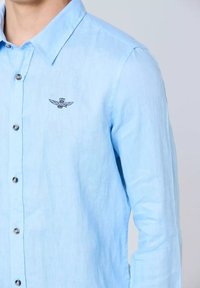 Camicia di lino azzurro chiaro con colletto a pullover, dotata di bottoni argentati e di un emblema ricamato nero con ali sul petto.