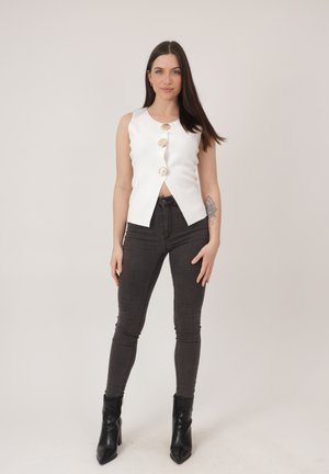 Kvinde med langt mørkt hår iført en hvid, ærmeløs top med knapper formet som muslingeskaller, sorte skinny jeans, sorte støvler og en blomster-tatovering på venstre underarm.